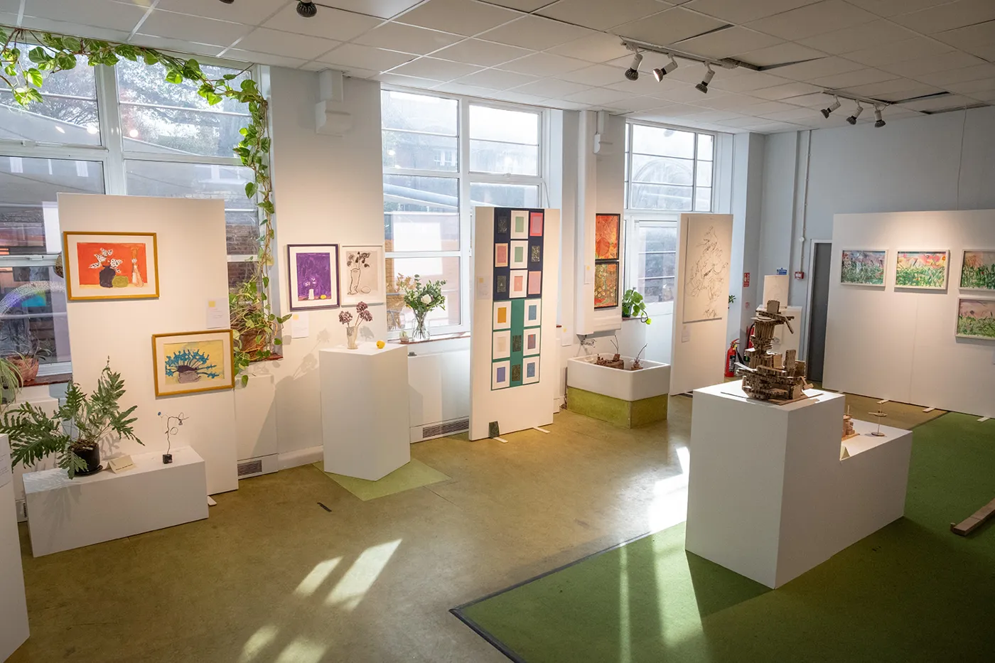 A bright room with a green floor showing paintings on white walls and sculptures on plinths